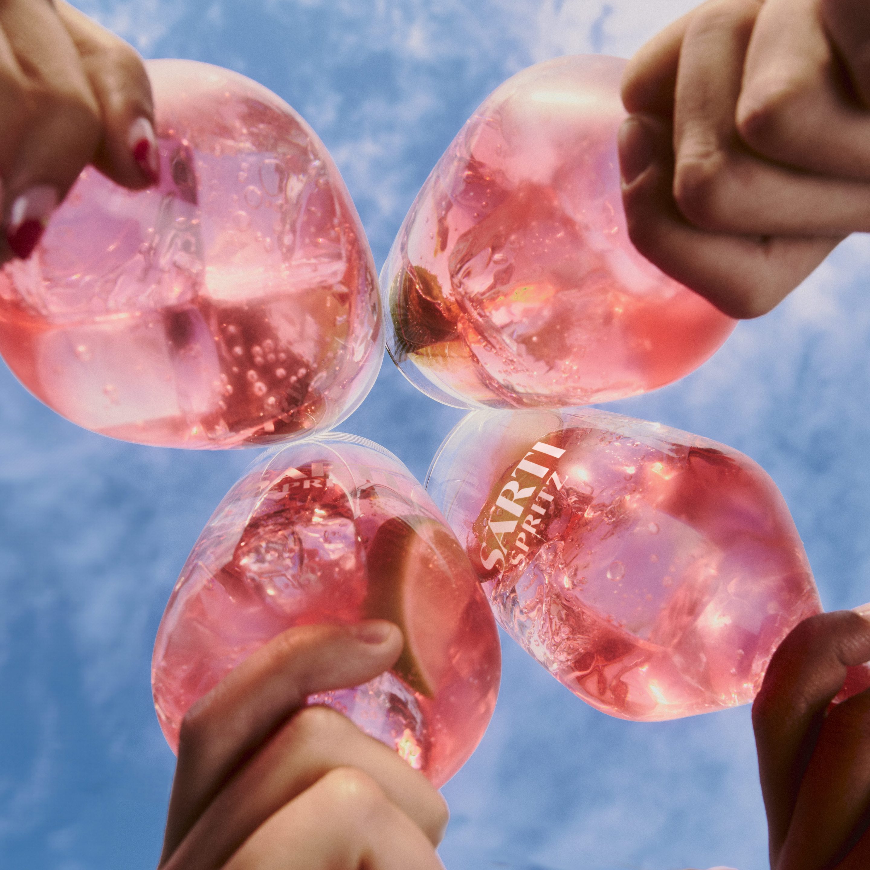 Four hands toast with pink Sarti Spritz cocktails against a blue sky.