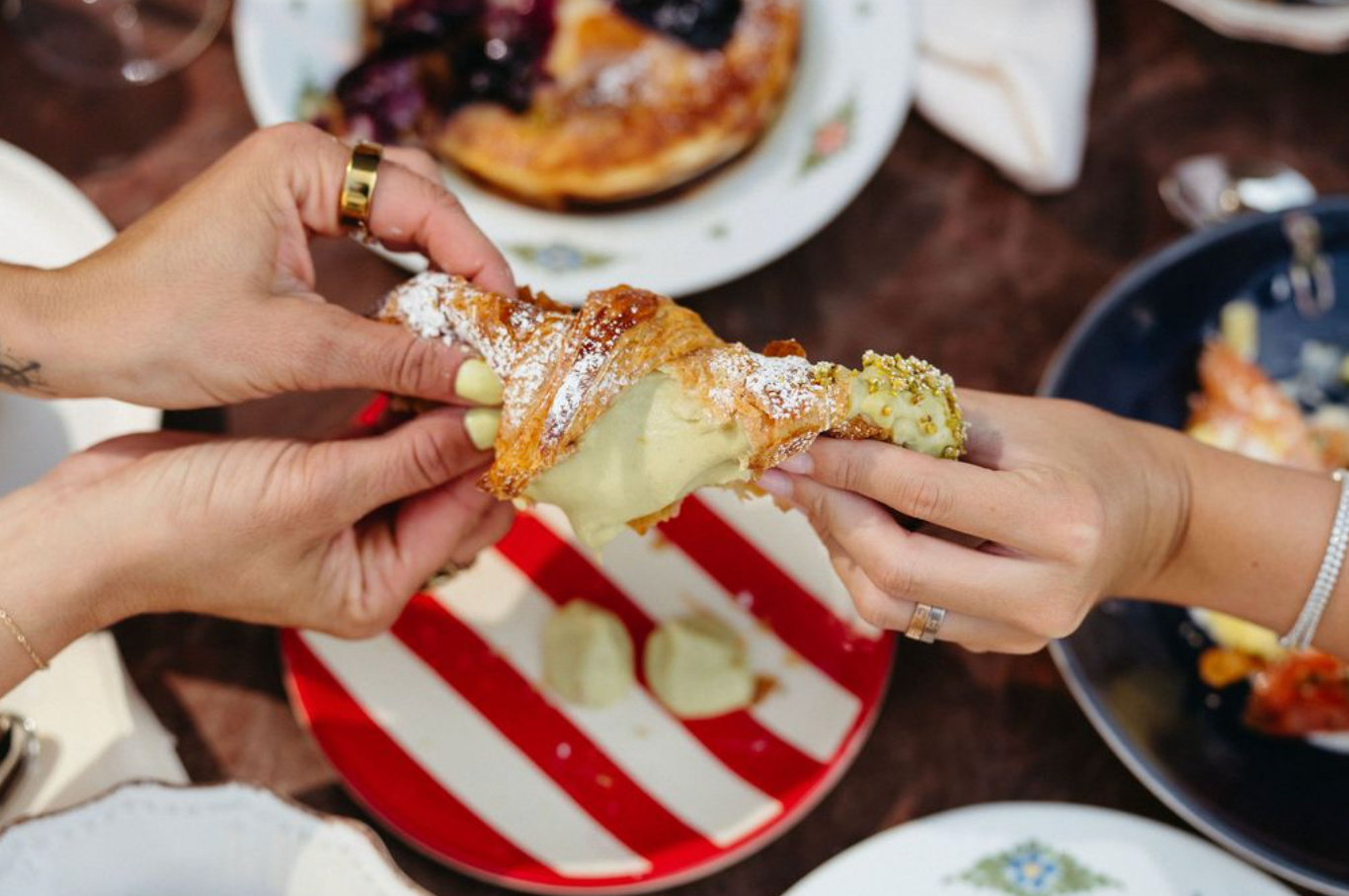 Two people pull apart a pistachio cream-filled cornetto over a red-and-white striped plate.