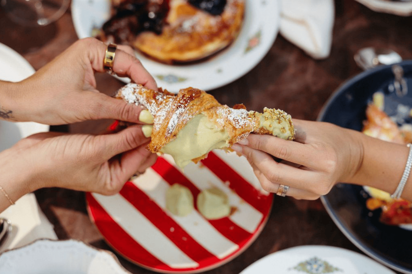 Two people pull apart a pistachio cream-filled cornetto over a red-and-white striped plate.