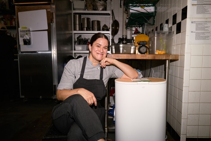 Chef Mary Attea seated beside a food recycler in a restaurant kitchen.