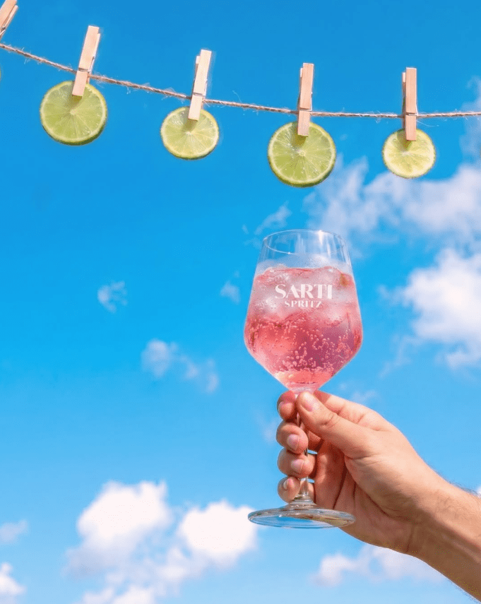 A hand holds a pink Sarti Spritz beneath lime slices hanging against a blue summer sky.