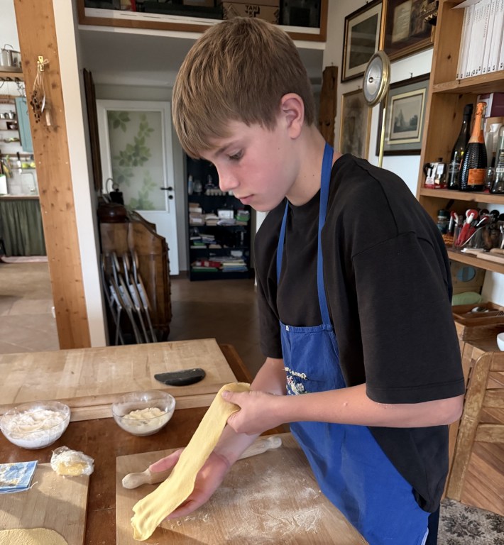 Boy wearing a blue apron holds a sheet of pasta dough over a floured board.