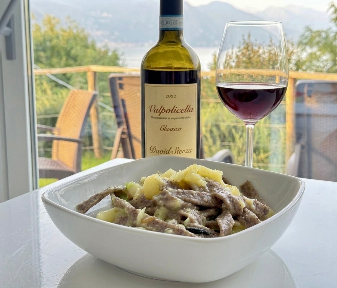 Bowl of pizzoccheri beside a bottle and glass of red wine with Lake Como in the background.