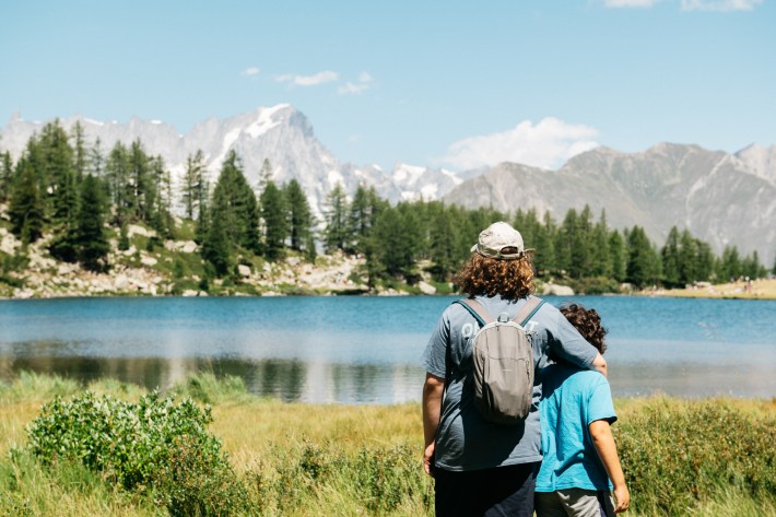 Visitors lounging on the grassy shore of Lago d’Arpy below larch trees and rocky slopes.