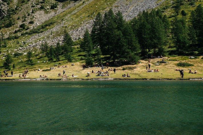 Visitors lounging on the grassy shore of Lago d’Arpy below larch trees and rocky slopes.