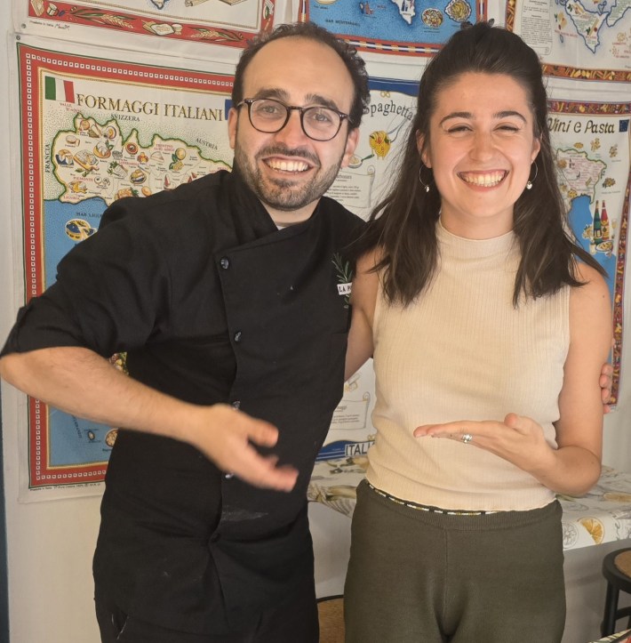 Luca Scaglia and Laura smiling in front of Italian food posters.