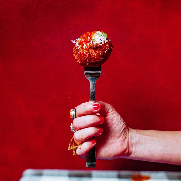A hand holds a fork with a meatball topped with red sauce, grated cheese and herbs against a red background.