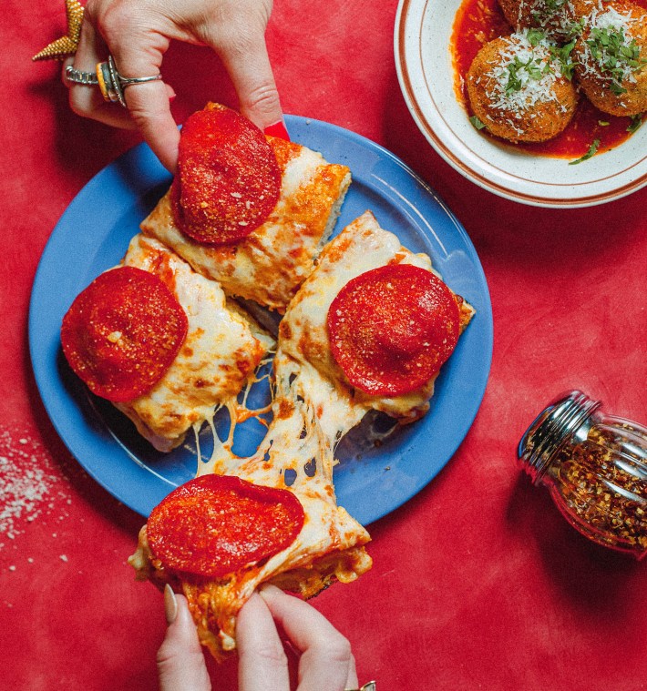 Hands pull apart pepperoni pizza squares with stretching cheese over a blue plate.