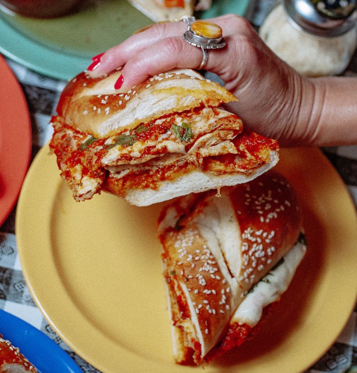 A hand holds a sandwich with breaded filling, red sauce and melted cheese on a sesame-topped roll.
