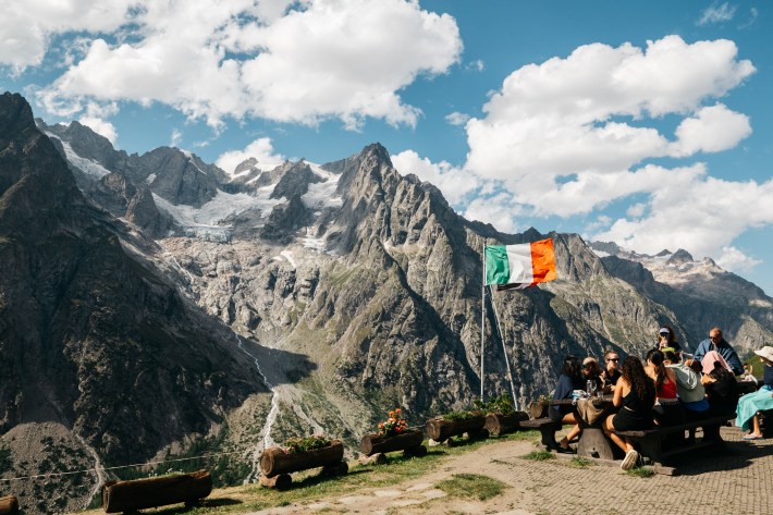Hikers sit at picnic tables beside an Italian flag at Rifugio Bonatti