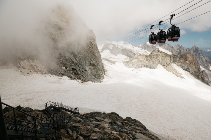 Three gondola cabins suspended above a glacier and jagged, cloud-covered peaks on Mont Blanc.