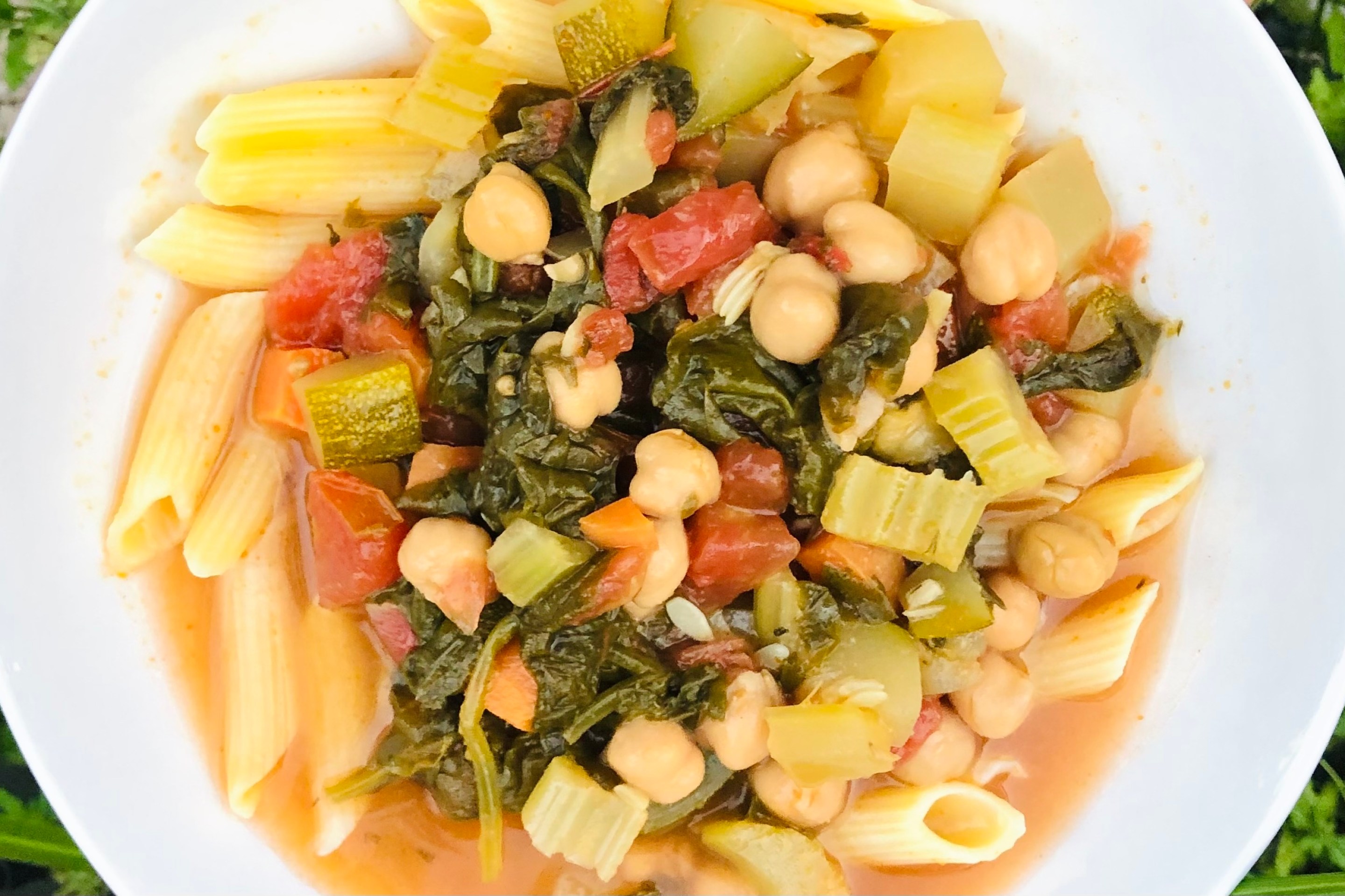 Vegetable soup with penne, chickpeas, tomatoes and leafy greens in a white bowl.