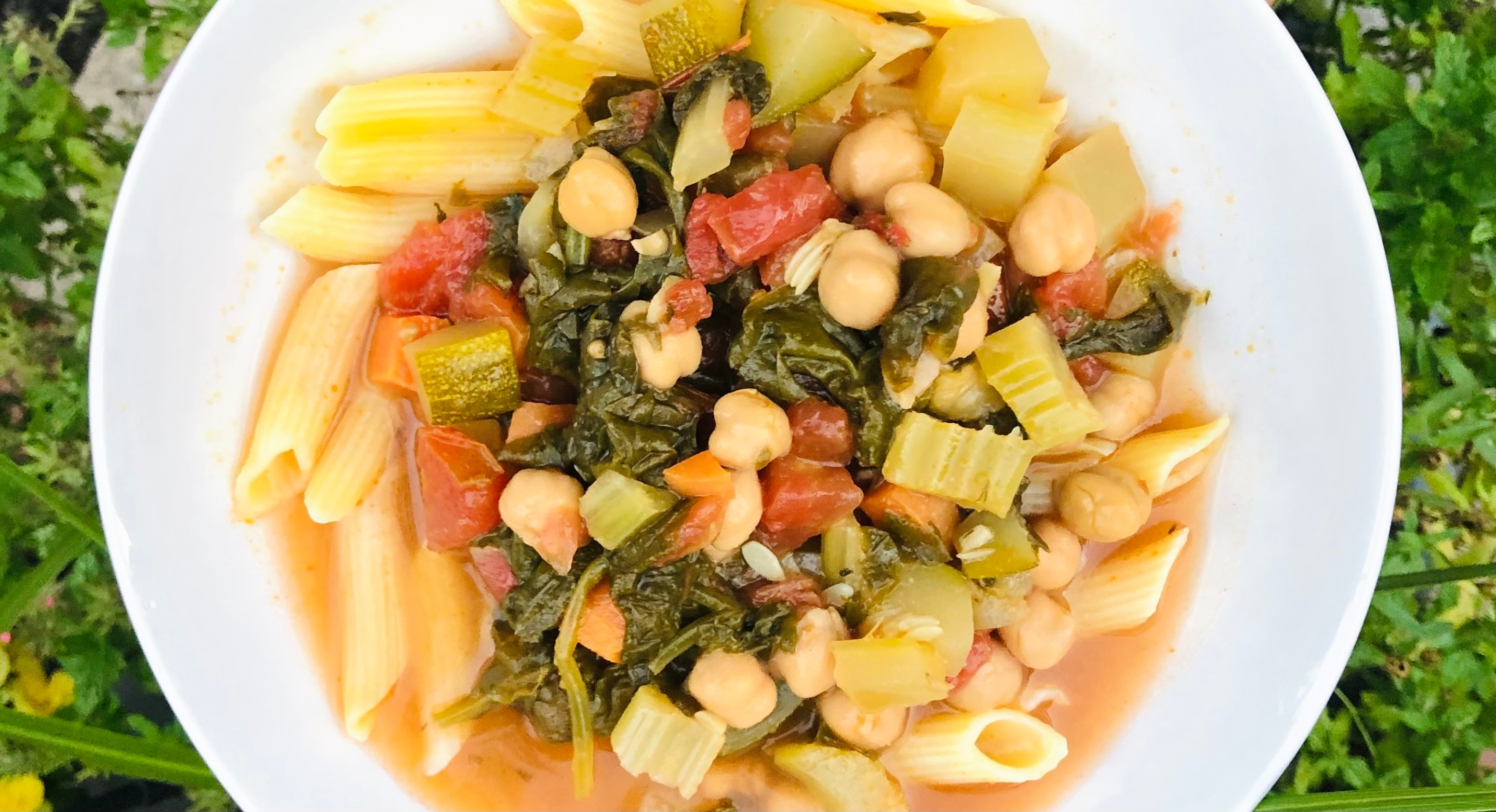 Vegetable soup with penne, chickpeas, tomatoes and leafy greens in a white bowl.