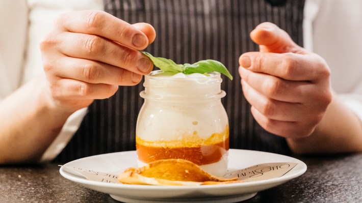 A person adds basil to Tapasotto’s egg in a glass, served in a jar with white and orange layers.