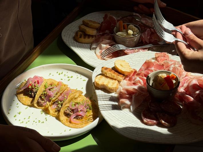 Beef tacos beside platters of cured meats, toasted bread and vegetables.