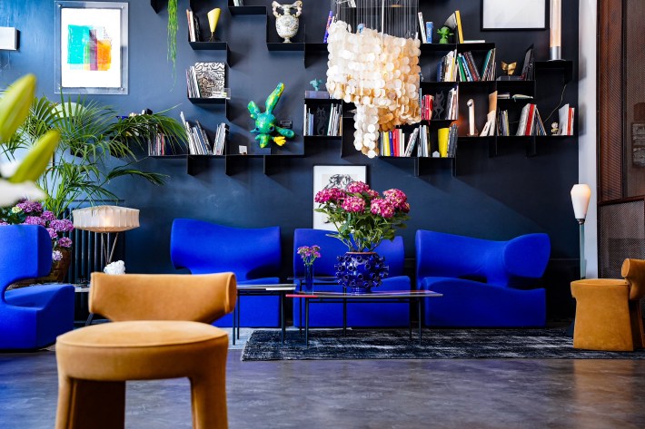 Hotel Trieste lounge with bright blue armchairs, bookshelves and a large hanging light above a glass table.