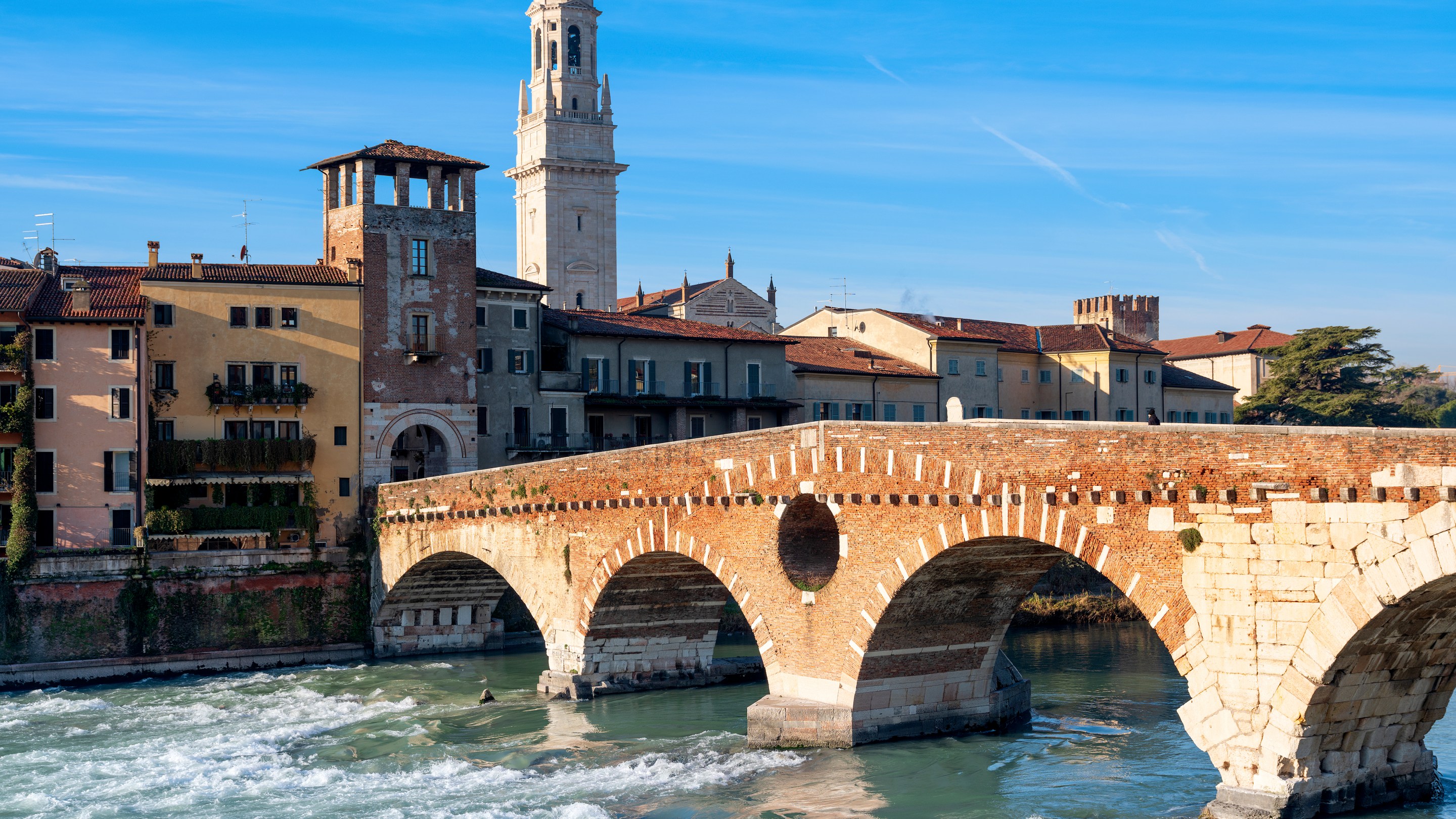 Ponte Pietra’s stone and brick arches cross the Adige River, with Verona’s buildings and a bell tower behind it.