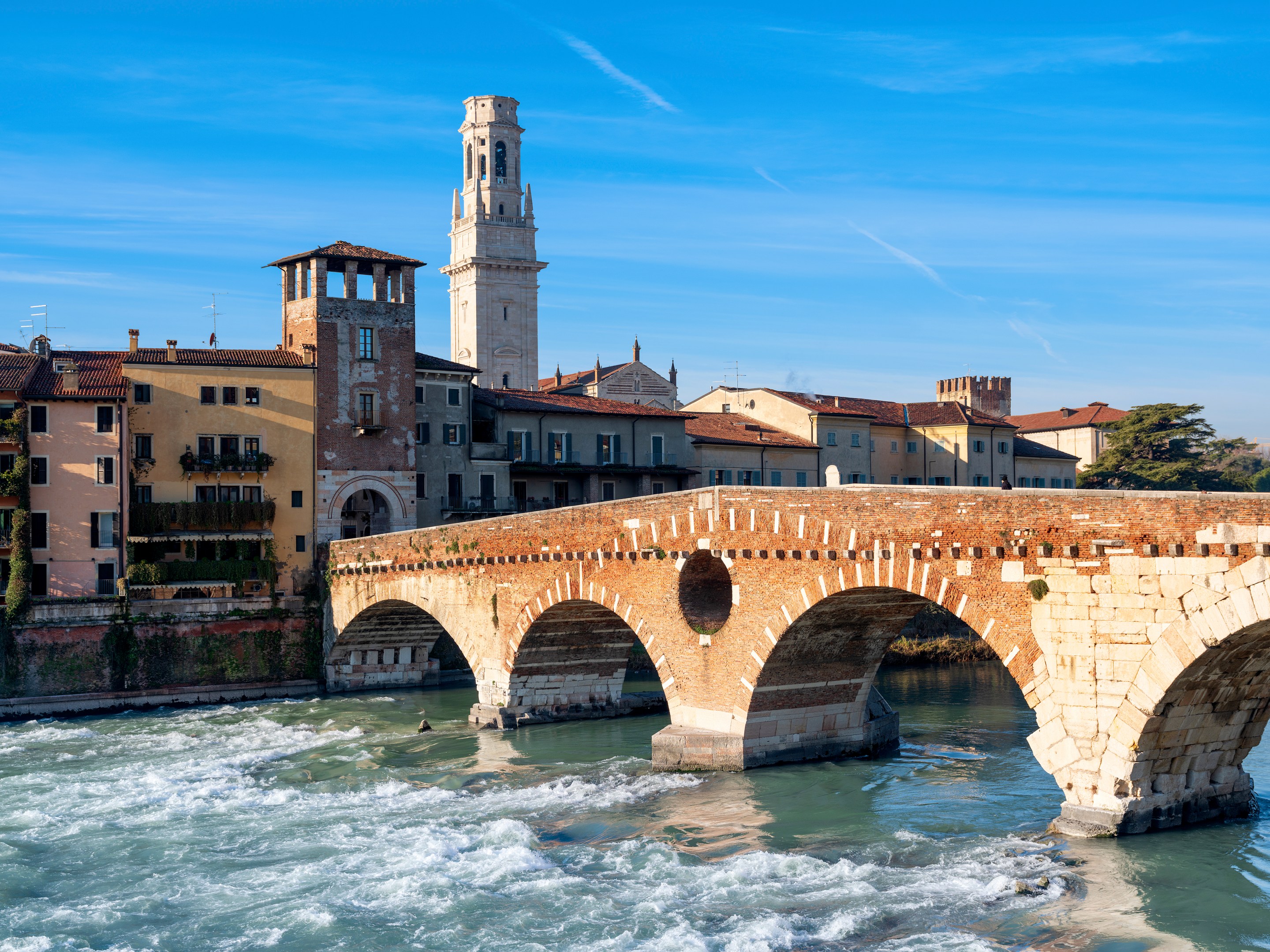 Ponte Pietra’s stone and brick arches cross the Adige River, with Verona’s buildings and a bell tower behind it.